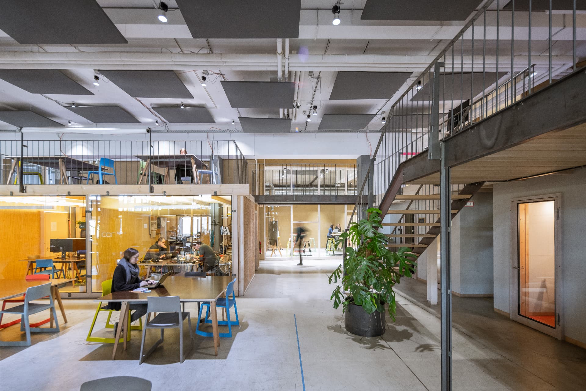 Heller, moderner Coworking-Space mit Glasbüros, Galerieebene und arbeitenden Personen an Tischen in einem offenen Raum.
